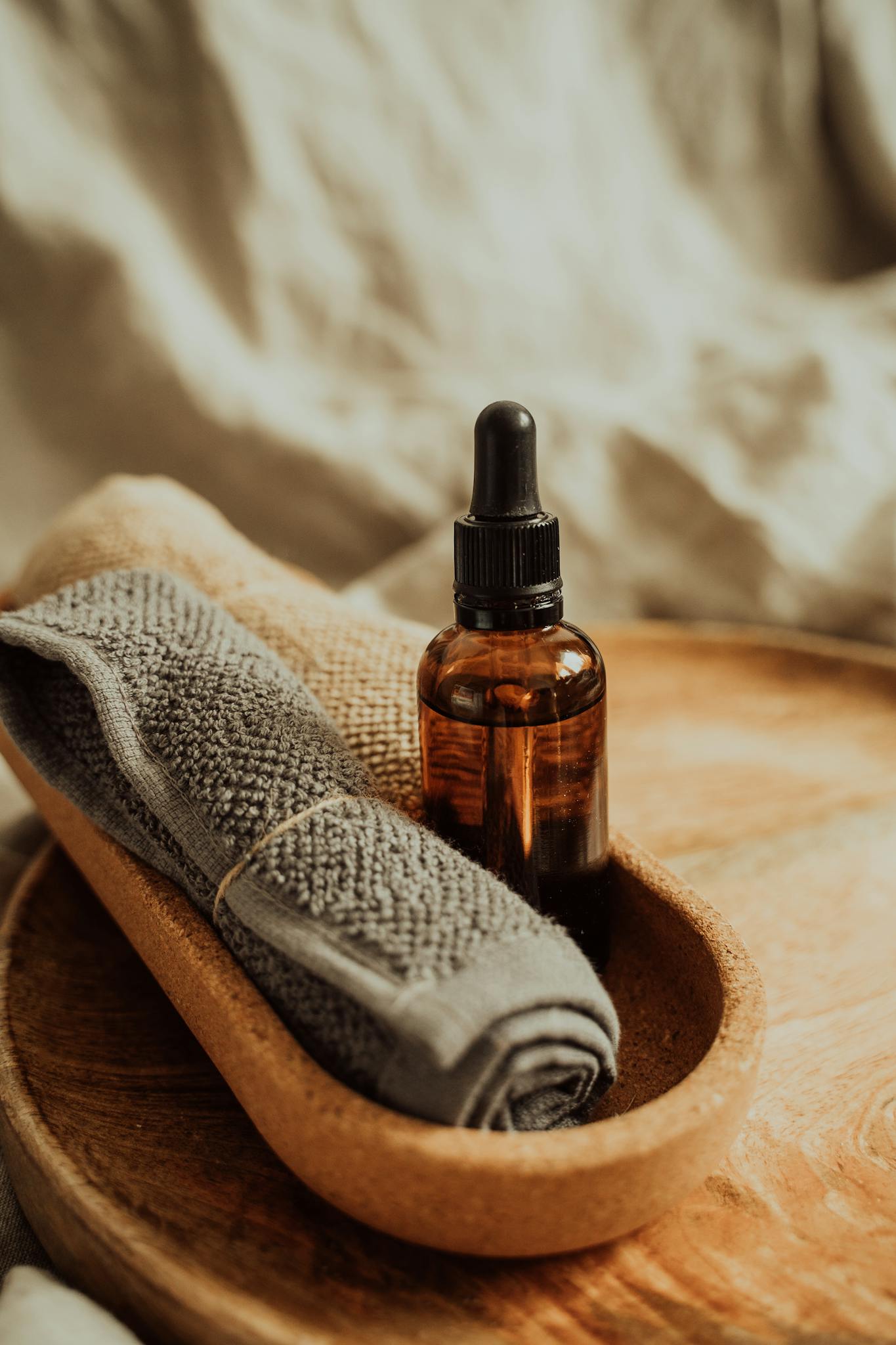 Close-up of a glass amber bottle with massage oil on a wooden tray, set with soft towels for a calming spa experience.