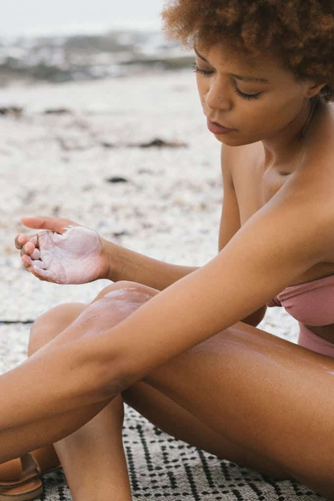 A woman applies sunscreen at the beach, embracing summer skincare and sun protection.