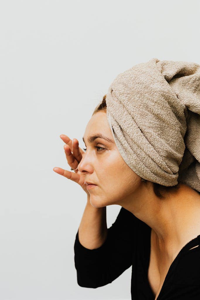Profile view of a woman applying skincare product, showcasing her beauty routine with a towel wrapped around her hair.